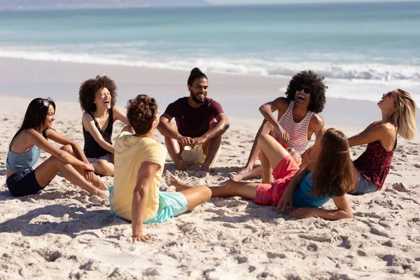 多种族的成年男女朋友们穿着短裤和T恤衫 坐在阳光灿烂的海滩上 一边聊天一边笑 背景是蓝天和大海 — 图库照片