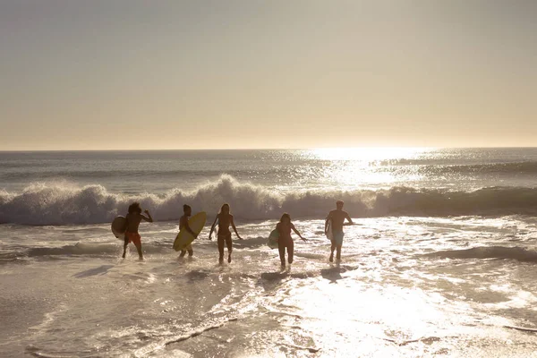 在海滩上度假时 一群多种族的男女朋友拿着冲浪板 冲向海浪 夕阳西下时的背影 — 图库照片