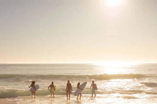 サーフボードを持ってビーチで休暇中の男性と女性の友人の多民族グループの背面ビューは 太陽が沈むと海に流れ込む — ストック写真