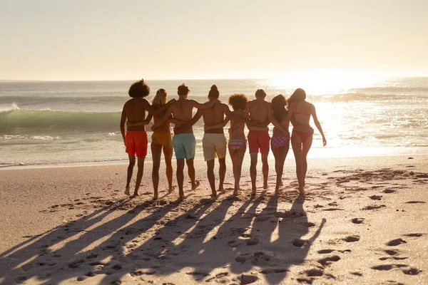 ビーチで楽しい休日の男性と女性の友人の多民族グループの背面ビューは 太陽が彼らの前に下るように海に向かって腕を組んで行の中を歩く — ストック写真