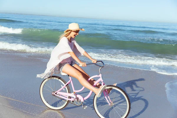 一个白种人女人 在阳光明媚的日子 戴着一顶帽子 骑着自行车 背景是大海 在海滩上享受时光 — 图库照片