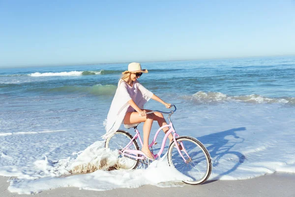 一个白种人女人 在阳光明媚的日子 戴着一顶帽子 骑着自行车 背景是大海 在海滩上享受时光 — 图库照片