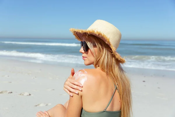 一个白种人女人 戴着帽子 在阳光灿烂的日子里 在海滩上享受时光 涂上防晒霜 并以大海为背景 — 图库照片