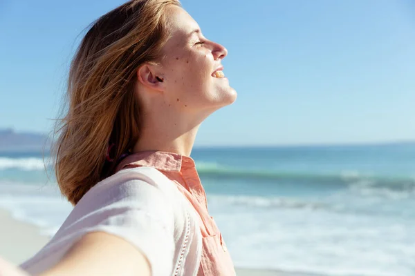 一个白种人女人在阳光明媚的日子里 在海滩上尽情享受时光 闭着眼睛 把手伸向镜头 身后是大海 — 图库照片