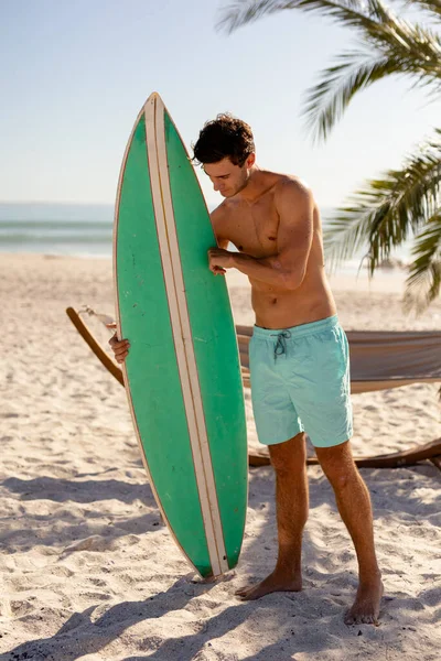 白人の男は 晴れた日にビーチで彼の時間を楽しんで 無表情に立ってサーフボードを保持し それを見て — ストック写真
