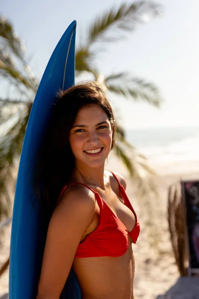 白人女性は晴れた日にビーチで彼女の時間を楽しんで 彼女の後ろにサーフボードを保持 カメラを見て — ストック写真