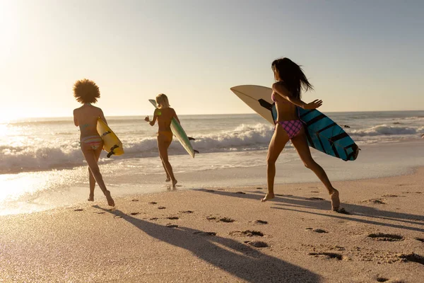 两个混血的种族和一个白人妇女在阳光灿烂的日子里 在海滩上享受着她们的时光 奔向大海 举着冲浪板 — 图库照片