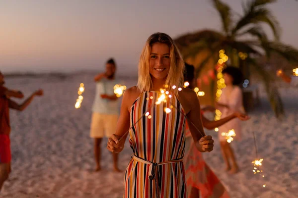 在夕阳西下的时候 白种人女人和她的朋友们一起在海滩上享受着她的时光 拿着火花 看着相机 和她的朋友们一起在后方微笑 — 图库照片