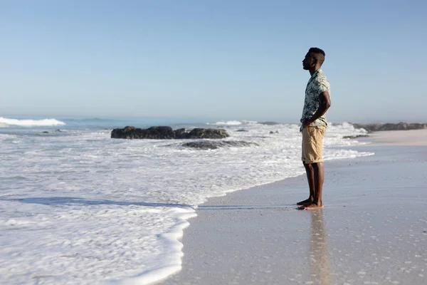 晴れた日にビーチで自由な時間を楽しんでいるアフリカ系アメリカ人の男性 ハワイのシャツを着て 砂の上に立って 景色を賞賛 — ストック写真