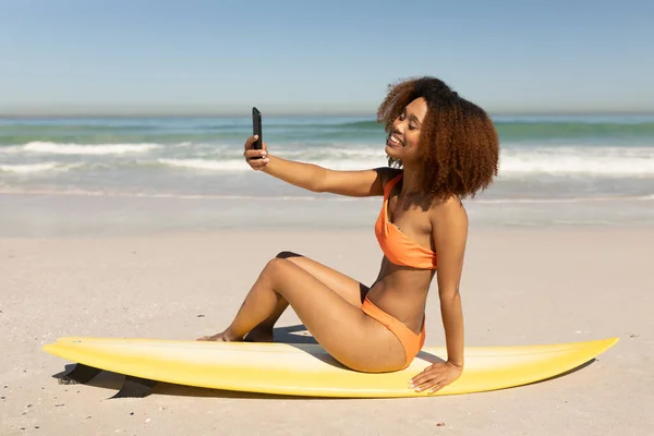 一个快乐 迷人的混血儿 在阳光灿烂的日子里尽情享受沙滩上的闲暇时光 面带微笑 玩得开心 坐在冲浪板上 拿着智能手机自拍 脸上闪烁着阳光 — 图库照片