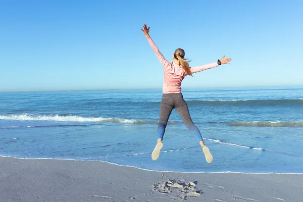 一个快乐的白人女人 穿着运动服 在阳光灿烂的日子里 在海滩上享受着快乐的时光 跳跃着面对大海 — 图库照片