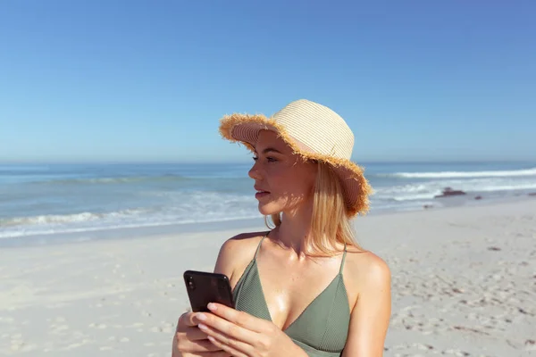晴れた日にビーチで時間を楽しむ魅力的なブロンドの白人女性 太陽の帽子を着用し スマートフォンを使用して 背景に青い空と海と 夏の熱帯のビーチ休暇 — ストック写真