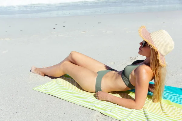 迷人的金发白人女人在阳光灿烂的日子里 在海滩上尽情享受时光 戴着帽子 躺在毛巾和日光浴上 背景是蓝天和大海 夏季热带海滩假期 — 图库照片