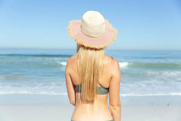 迷人的金发白人女人在阳光灿烂的日子里 在海滩上享受着时光 戴着太阳帽 面对着大海 背景是蓝天和大海 夏季热带海滩假期 — 图库照片