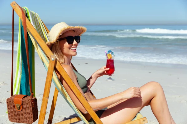 晴れた日にビーチで時間を楽しむ魅力的なブロンドの白人女性 デッキチェアに座って カクテルを保持 背景に青い空と海と 夏の熱帯のビーチ休暇 — ストック写真