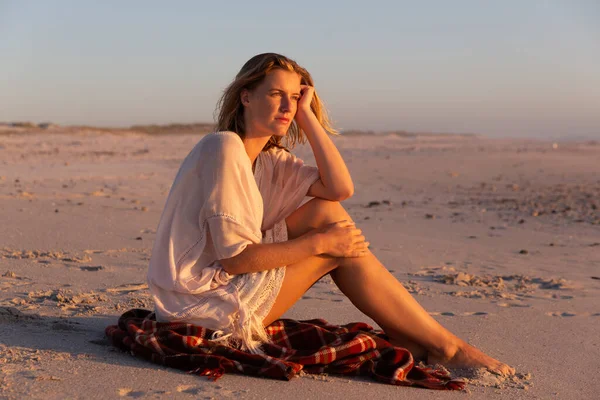 魅力的なブロンドの白人女性は 日没時にビーチで時間を楽しんで 毛布の上に座って遠くを見て 背景に夕日の空と海があります 夏の熱帯のビーチ休暇 — ストック写真