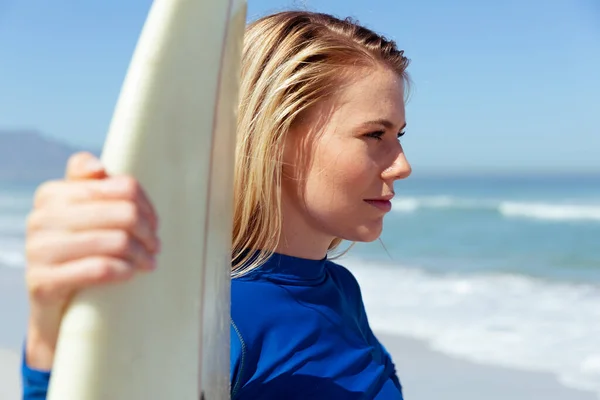 迷人的金发白人女人 在阳光灿烂的日子里 在海滩上享受着时光 手握冲浪板 面带微笑 背景是蓝蓝的天空和大海 夏季热带海滩假期 — 图库照片