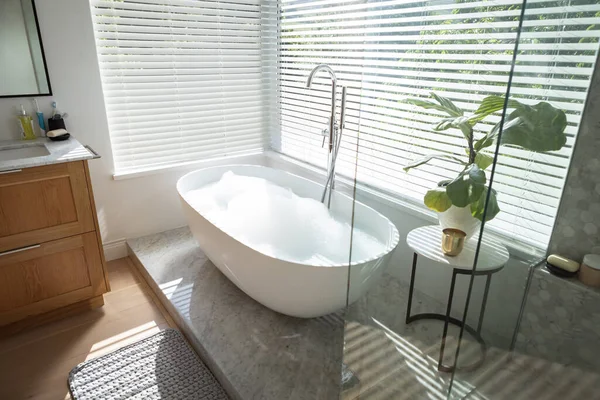 现代化的浴室内部有免费站立的浴室 窗户很大 外面有绿树和植物 有白色浴缸和银色水龙头 灰色淋浴和有植物的桌子 — 图库照片