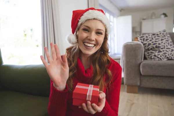 自宅で時間を過ごす白人女性の肖像画 リビングルームで 笑みを浮かべて クリスマスの帽子をかぶって 手を振ってギフトボックスを保持 Covid 19コロナウイルス隔離ロックダウン中の社会的距離 — ストック写真