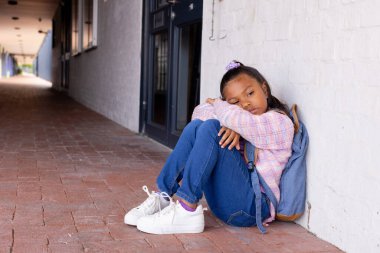 Biracial girl appears sad, sitting alone against a wall with copy space in school. Her downcast eyes and slumped posture suggest loneliness in an outdoor setting. clipart