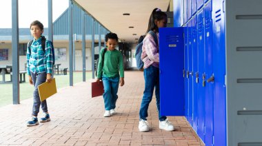 Biracial boy and African American boy walk past a biracial girl at school lockers. The boys carry folders, suggesting a break between classes, while the girl examines her locker. clipart
