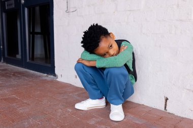 African American boy sits on the ground at school, looking sad. Dressed in casual attire, he rests against a white wall outdoors, conveying a mood of contemplation. clipart