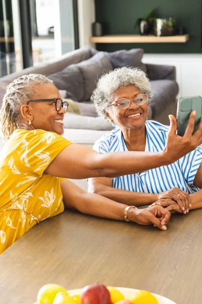 Senior African American woman and senior biracial woman are smiling while taking a selfie at home. Dressed casually, they share a moment of joy in a modern kitchen setting.