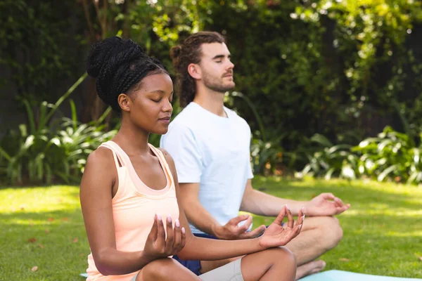 Diverse couple, a young African American woman and Caucasian man, meditate in a serene garden. Both are in lotus position, embracing tranquility and mindfulness outdoors.