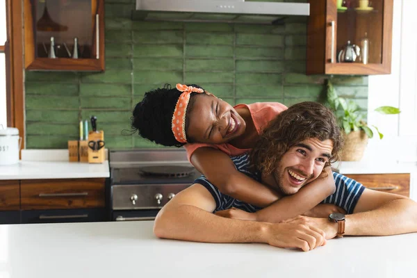 Diverse couple young African American woman embraces a young Caucasian man from behind in a kitchen. Both are smiling, conveying a sense of joy and companionship in a domestic setting.