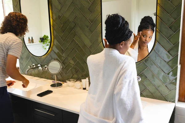Diverse couple: Young African American woman applies makeup in a bathroom mirror. She wears a white robe, creating a relaxed, personal grooming atmosphere.