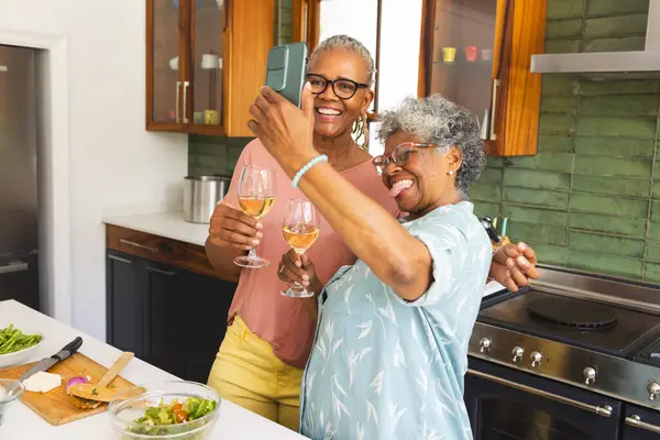 Senior African American woman and senior biracial woman share a joyful toast in a kitchen. Holding wine glasses, they celebrate with a selfie, surrounded by cooking ingredients and a modern stove.