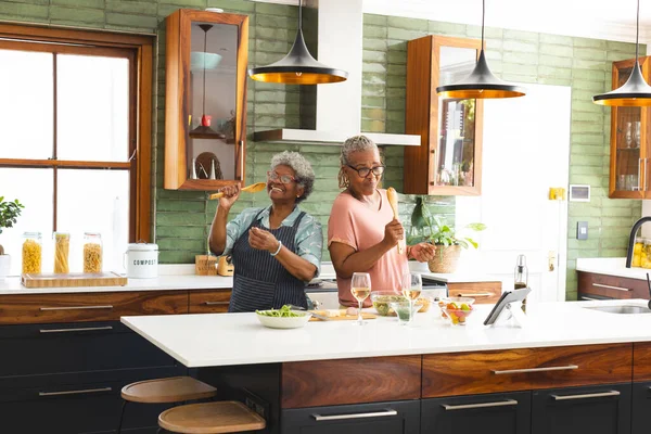 Senior African American woman and senior biracial woman are cooking together in a modern kitchen. They share a joyful moment, dancing lightly with wine glasses nearby.