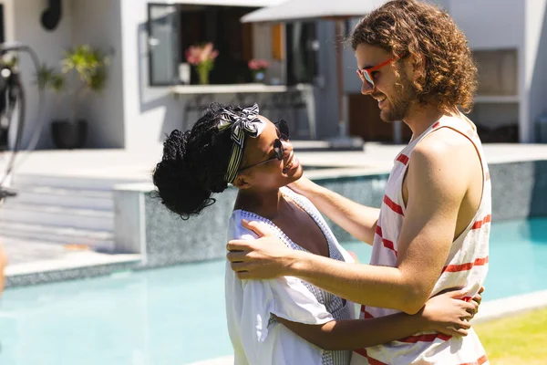 Diverse couple: Young African American woman and Caucasian man embrace by a pool. She wears a headscarf and sunglasses; he sports curly hair and red-striped tank top.