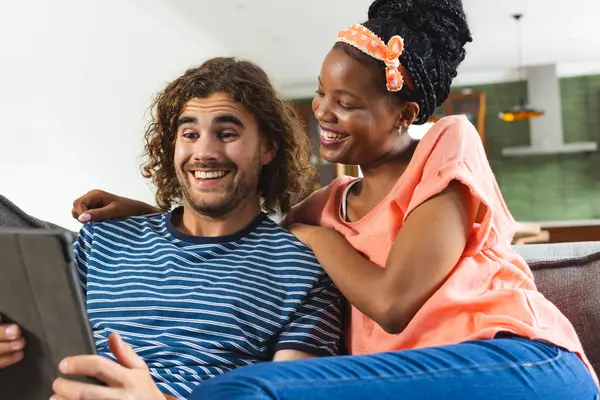 Diverse couple young African American woman and young Caucasian man share a tablet, smiling together. They are seated comfortably indoors, suggesting a moment of leisure and connection.