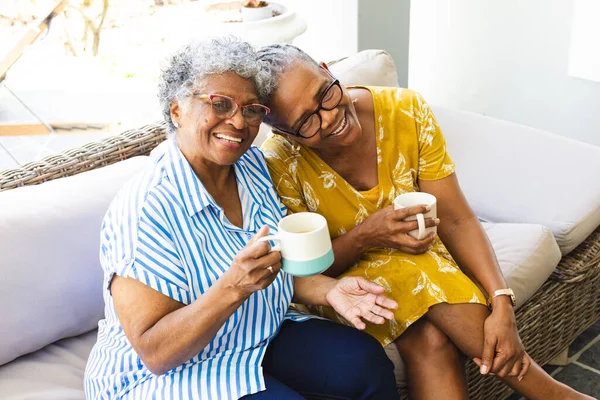 Senior African American woman and senior biracial woman share a joyful moment with coffee at home. They are sitting closely, exuding warmth and companionship in a bright, homey setting.