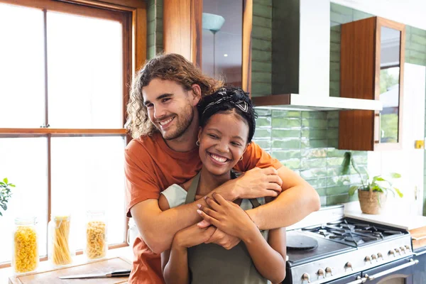 Diverse couple young Caucasian man embraces a young African American woman in a sunny kitchen. Both are smiling, wearing casual clothes, conveying a sense of warmth and affection.