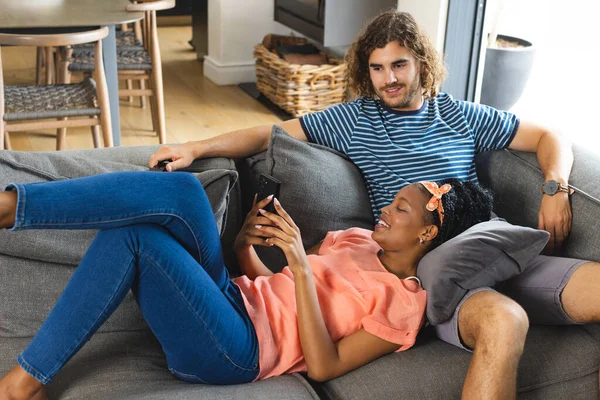 Diverse couple young African American woman and Caucasian man relax on a sofa, browsing a smartphone. She wears an orange top and headband; he sports a striped shirt, both enjoying a casual day at home.