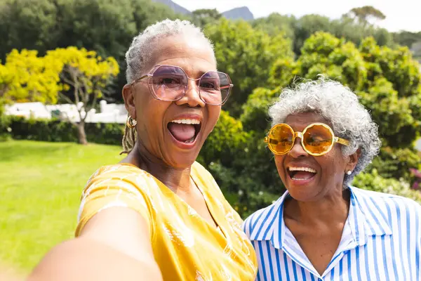 Senior African American woman and senior biracial woman share a joyful moment outdoors. Both are wearing vibrant glasses and bright smiles, capturing a sense of friendship and happiness.