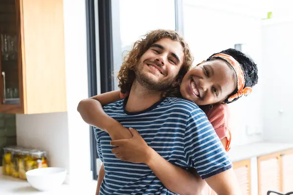 Diverse couple young African American woman embraces a young Caucasian man from behind, both smiling joyfully. They are in a bright kitchen setting, suggesting a close and comfortable relationship.