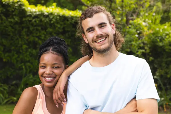 Diverse couple, a young African American woman and young Caucasian man, are smiling outdoors. They stand close together, suggesting a friendly or intimate relationship, amidst greenery.