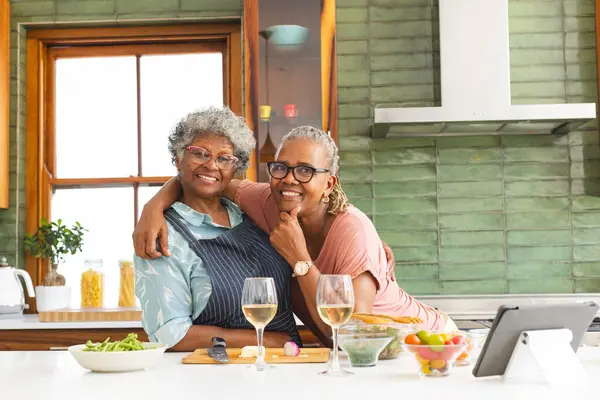Senior African American woman and senior biracial woman share a warm embrace in a kitchen. They are surrounded by a salad, wine glasses, and a tablet, suggesting a cozy meal preparation or a cooking session.