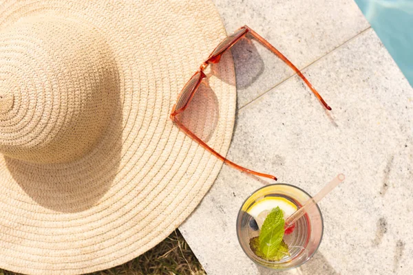 A straw hat, sunglasses, and a refreshing drink sit under the sunlight with copy space. The scene suggests a relaxing day at an outdoor setting, a beach or poolside.