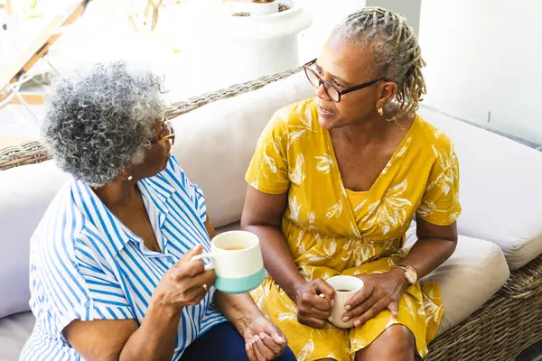 Senior African American woman and senior biracial woman enjoy a conversation over coffee at home. Both are dressed casually, creating a relaxed atmosphere in a bright, home setting.