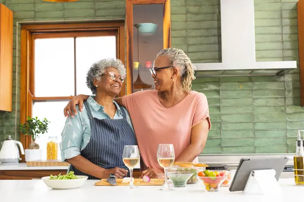 Senior African American woman and senior biracial woman share a warm moment in a sunny kitchen. They stand close, smiling, with wine glasses on the counter, suggesting a relaxed, intimate gathering.