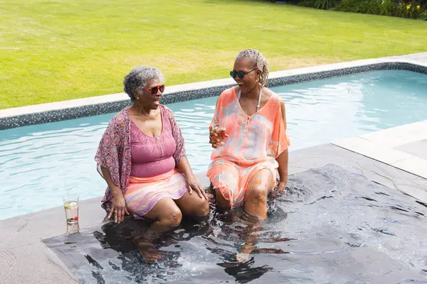 Senior African American woman and senior biracial woman enjoy a poolside chat. Both are dressed in swimwear with cover-ups, embodying leisure and friendship.