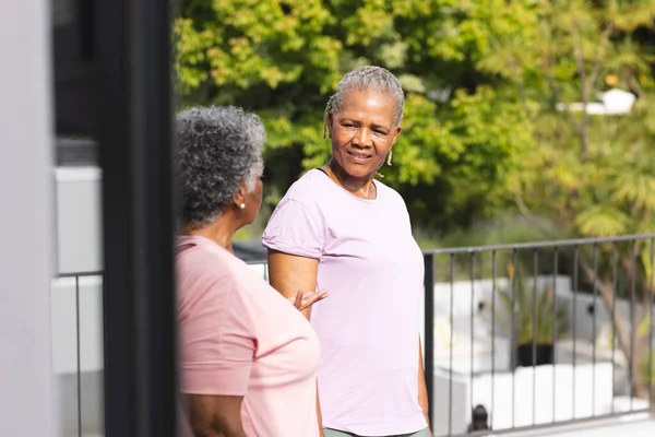 Senior African American woman converses with a senior biracial woman on a sunny balcony. Both are dressed casually, enjoying a friendly chat outdoors.