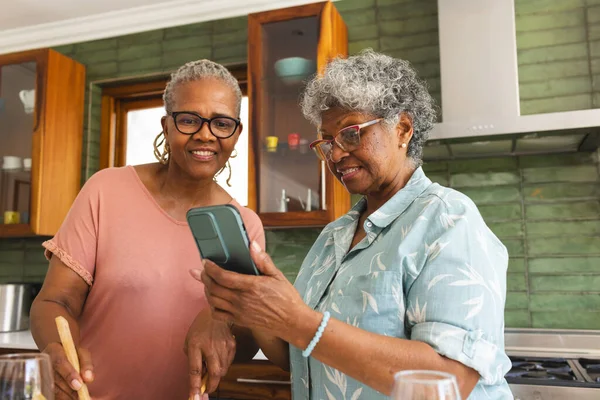 Senior African American woman and senior biracial woman share a moment in a kitchen. They are smiling while looking at a smartphone, surrounded by cooking utensils.