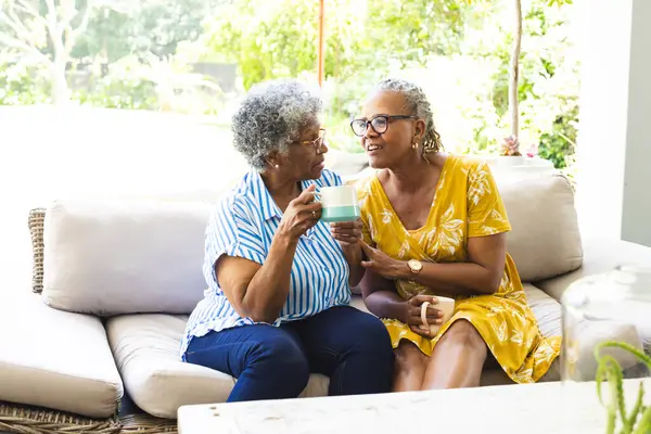 Senior African American woman and senior biracial woman share a moment over a cup of tea at home. They are seated comfortably on a couch, enjoying each other\'s company in a bright, homey setting.