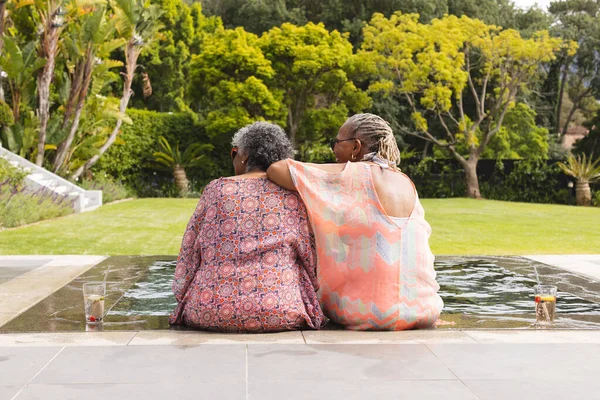 Senior African American woman and senior biracial woman sit close, overlooking a garden. They share a moment of relaxation and companionship by a reflective pool, surrounded by lush greenery.
