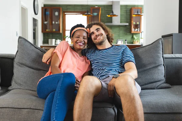 Diverse couple young African American woman and Caucasian man sit closely on a sofa, smiling warmly. Their casual attire and cozy home setting suggest a relaxed and affectionate moment together.